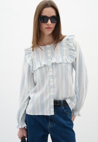 Blusa leggera a maniche lunghe con righe blu e bianche, caratterizzata da dettagli arricciati sul davanti e un design con bottoni. Indossata con jeans scuri.