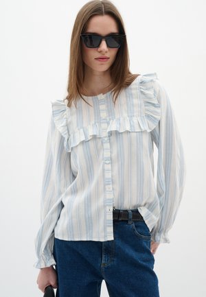 Blouse légère à manches longues avec des rayures bleues et blanches, présentant des détails froncés à l'avant et un design à boutons. Portée avec un jean noir.