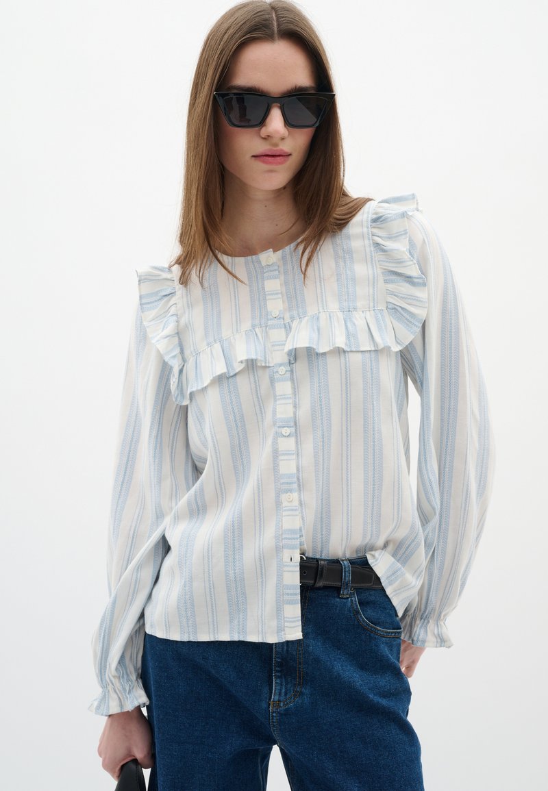 Blusa leggera a maniche lunghe con righe blu e bianche, caratterizzata da dettagli arricciati sul davanti e un design con bottoni. Indossata con jeans scuri.