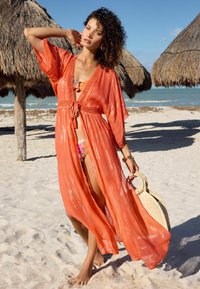 Cubierta fluida naranja con tela transparente y acentos brillantes, que presenta un lazo en la cintura y mangas amplias, combinada con un bikini multicolor.