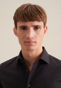 Giovane uomo con capelli castani corti che indossa una camicia nera con bottoni, rivolto verso un fondale beige uniforme.