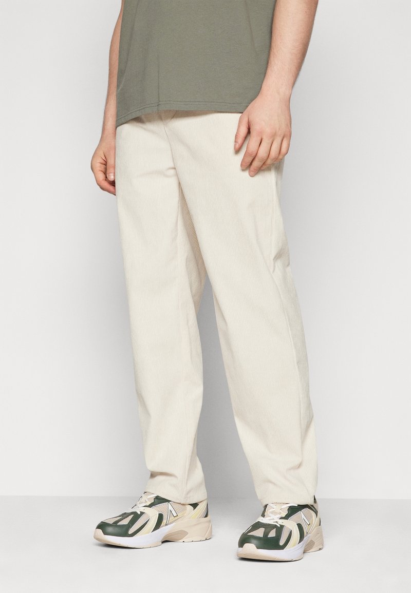 Personne portant un pantalon large en velours côtelé beige, des baskets vertes et blanches et une chemise vert olive, debout devant un fond uni.