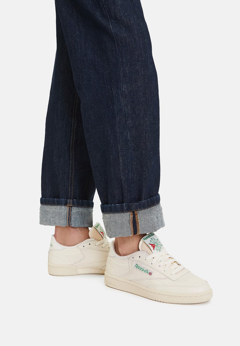 Osoba nosiaca tmavomodré rifle s manžetami a krémové tenisky Reebok s detailmi v zelenej a červenej farbe na bielom pozadí.