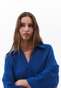 Camicia button-up blu, strutturata, con colletto e maniche lunghe. Il tessuto presenta un leggero motivo a griglia. Il modello ha le braccia incrociate.