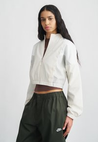 Κοντό μπεζ σακάκι με ψηλό γιακά και ελαστικό τελείωμα, με γκρι πλαϊνά πάνελ. Φορεμένο πάνω από καφέ τοπ και πράσινο παντελόνι.