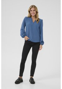 Blaue Langarmbluse mit V-Ausschnitt und gerafften Manschetten, kombiniert mit schwarzen Skinny-Jeans und schwarzen Plateauschuhen. Glatte Textur.