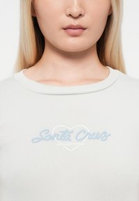 Tricou gri deschis cu dungi, însoțit de un text brodat albastru "Santa Cruz" în interiorul unui design în formă de inimă. Decupaj rotund, textură moale.