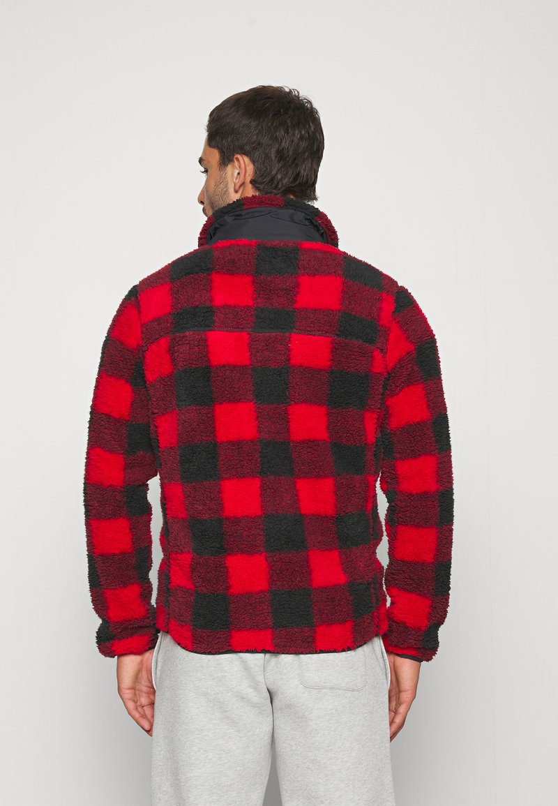 Veste en polaire à carreaux rouge et noir avec un col haut, surface texturée et poignets élastiques, portée avec un pantalon de survêtement gris.