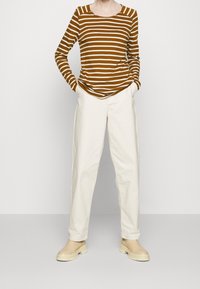 Striped long-sleeve shirt in brown and cream, paired with cream trousers and beige shoes. Material appears soft, with relaxed fit and round neckline.
