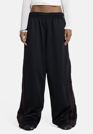 Oseba, oblečena v široke črne hlače z rdečimi stranskimi črtami, beli kratki dolg rokav, in črne čevlje, stoji ob belem ozadju.