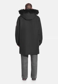 Czarna parka z kapturem obszytym futrem, o prostym kroju, z długimi rękawami i subtelną naszywką z logo na lewym ramieniu. W zestawie z szarymi spodniami.