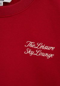 Tissu rouge avec texte blanc brodé ""The Leisure Sky Lounge"" près de l'encolure d'un vêtement.