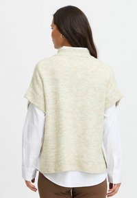 Rövidujjú, világos bézs pulóver, magas gallérral, puha textúrával és enyhén oversized fazonnal, fehér ing fölé rétegezve.