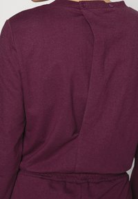 Top bordeaux caratterizzato da una finitura texturizzata, chiusura sul retro con bottoni e dettaglio in vita arricciato per una forma aderente.