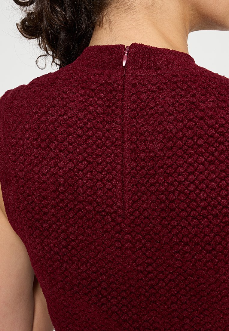 Top de tela texturizada burdeos con un sutil patrón en relieve, con un detalle de cremallera en la parte posterior y un diseño de cuello alto.
