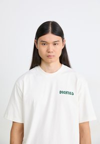 Hvit bomull t-skjorte med korte ermer, med grønn tekst "DICKIES" på venstre brystområde. Glatt tekstur, klassisk passform.