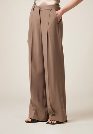 Brązowe spodnie o szerokich nogawkach noszone przez osobę z jedną ręką w kieszeni i czarnymi sandałami na palce na jasnym tle.