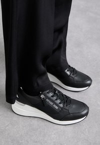 Sorte sneakers lavet af en blanding af læder og mesh-materialer, med en hvid gummisål, sorte snørebånd og en metallisk detalje på hælen.