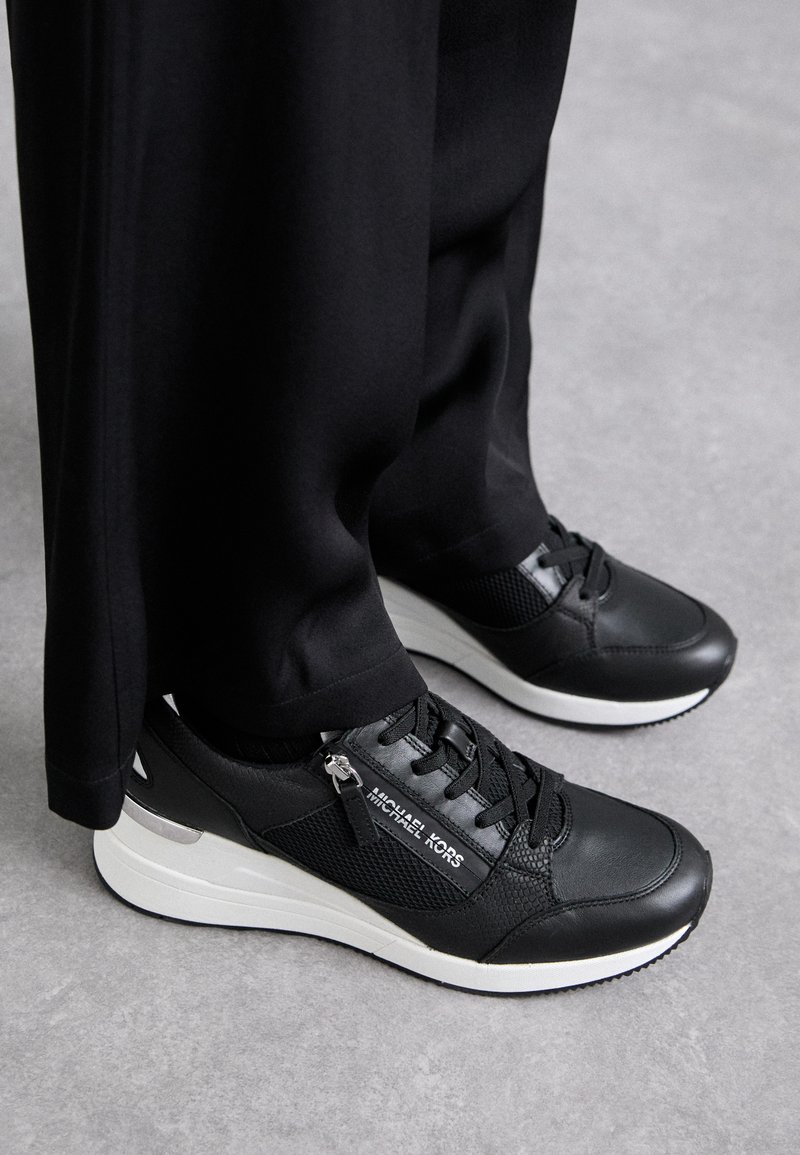 Sorte sneakers lavet af en blanding af læder og mesh-materialer, med en hvid gummisål, sorte snørebånd og en metallisk detalje på hælen.