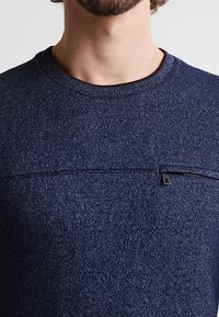 Uomo che indossa un maglione a collo rotondo di un blu scuro con una texture, caratterizzato da un dettaglio con zip orizzontale sul petto.