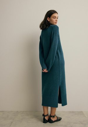 Lang, mørk teal strikket kjole med en afslappet pasform, ribbede manchetter, sideslids og blød tekstur. Sammen med sorte flade sandaler med stropper.