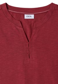 Chemise rouge avec une petite fente en V au col et trois fines ouvertures horizontales cousues sous le col avec une étiquette blanche indiquant CECIL.