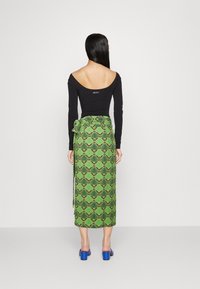 Schwarzes langärmliges Oberteil mit offenem Rücken, kombiniert mit einem grünen gemusterten Rock mit Blattdesign, der an der Taille gebunden ist. Blaue Blockabsatzschuhe.