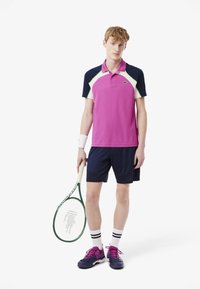 Rosa pikétröja med marinblå axlar och gröna detaljer, ihop med mörka shorts. Tillbehör inkluderar ett vitt handledsband och en tennisracket.