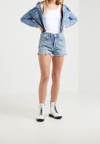 Chaqueta de mezclilla, camiseta sin mangas blanca y pantalones cortos de mezclilla azul deshilachados. Completado con botas de combate blancas con cordones. El fondo es blanco liso.