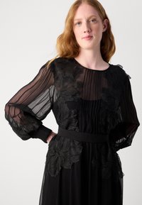 Robe noire avec des manches longues plissées et transparentes, des appliqués floraux texturés, une ceinture ajustée à la taille et une jupe fluide.