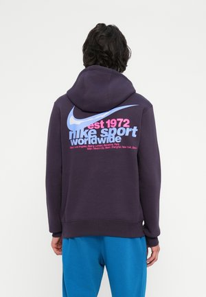 Persoon draagt een donkere hoodie met een grote blauwe Nike-swoosh en roze en blauwe tekst op de achterkant met de woorden "est 1972 nike sport worldwide".