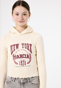 Sudadera corta beige con un estampado frontal que dice "NEW YORK GARCIA 1972", con un dobladillo en forma de concha y capucha adjunta.