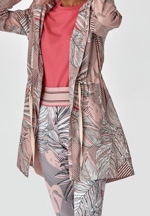 Femme portant une veste imprimé tropical rose et blanc et un pantalon assorti sur un t-shirt rose uni, ajustant la capuche de la veste.