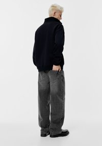 Giacca in pile nera con collo alto, abbinata a jeans a gamba larga grigi a righe e calzature nere, vista da dietro.