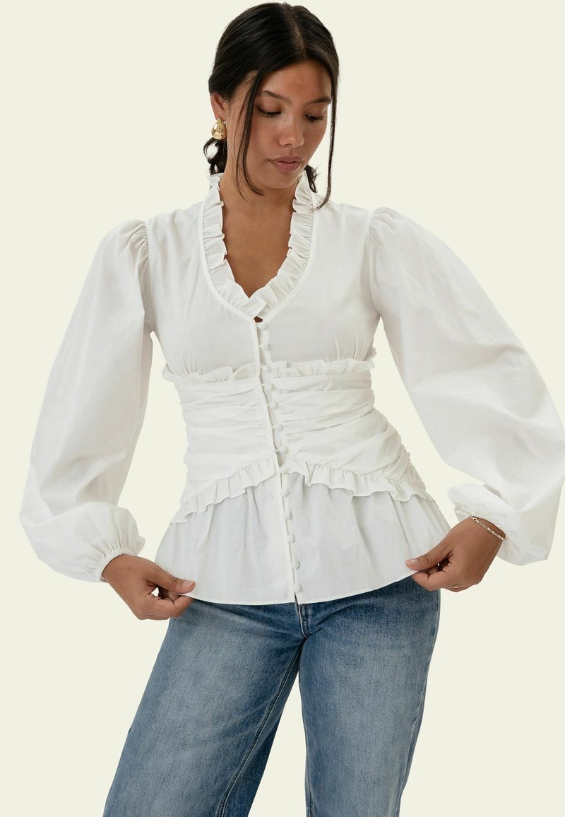 Femme ajustant un chemisier blanc à manches longues avec des volants et des boutons, portant un jean bleu et des boucles d'oreilles en or devant un fond uni.
