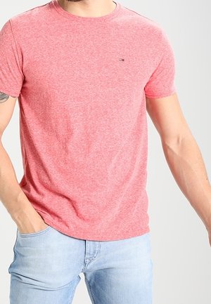 Mężczyzna w różowym t-shircie z krótkim rękawem i jasnoniebieskich dżinsach, stoi z jedną ręką w kieszeni na jednolitym tle.