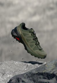 Scarpa da trail running Salomon verde e nera con suola robusta, sospesa nell'aria sopra un terreno roccioso con piccole particelle di terra che cadono.