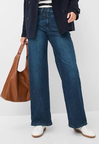 Jeans in denim scuro a gamba larga con un aspetto leggermente sbiadito. Abbinati a sneakers bianche e a una borsa tote in pelle marrone. Top a righe visibile.