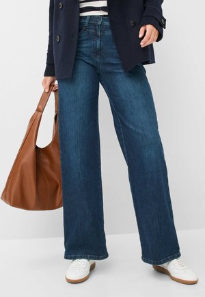 Jeans in denim scuro a gamba larga con un aspetto leggermente sbiadito. Abbinati a sneakers bianche e a una borsa tote in pelle marrone. Top a righe visibile.