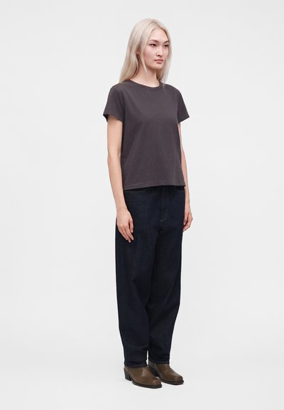 Jeune personne aux longs cheveux clairs portant un t-shirt gris foncé, un jean large sombre et des bottes marron, debout devant un fond uni.