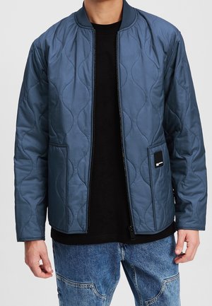 Mann, der eine gesteppte blaue Bomberjacke über einem schwarzen Shirt und blauen Jeans trägt, steht mit entspannten Armen an den Seiten.