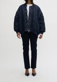 Granatowa pikowana kurtka bomberka z zamkiem błyskawicznym i zatrzaskami, z prążkowanymi mankietami i luźnym krojem, zestawiona z ciemnymi dżinsami i czarnymi szpilkami.