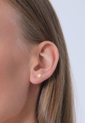 Boucle d'oreille en or avec une surface texturée, accrochée à la partie inférieure de l'oreille. Les cheveux environnants sont raides et légèrement ondulés, révélant la boucle d'oreille.
