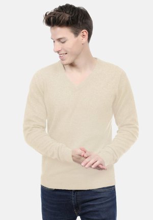 Pull V en col en beige clair en matière douce, avec des poignets côtelés, associé à un jean bleu foncé. Design épuré, idéal pour un style décontracté.