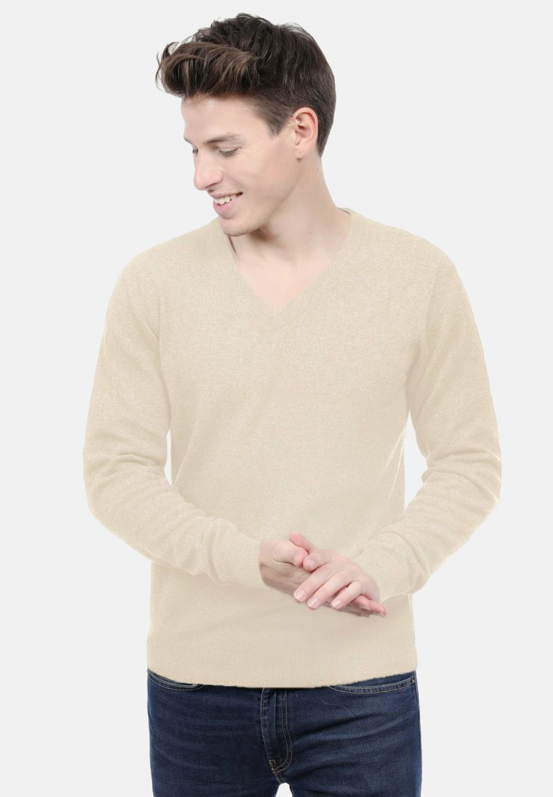Pull V en col en beige clair en matière douce, avec des poignets côtelés, associé à un jean bleu foncé. Design épuré, idéal pour un style décontracté.