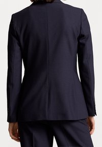 Kvinna i en figurnära mörkblå kavaj och matchande byxor, sedd bakifrån mot en enkel vit bakgrund.