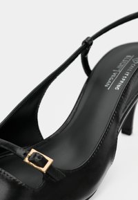Černý lodičkový bot s vysokým podpatkem, se slingback páskem a zlatou sponou, se špičatou špičkou a označením "Calf Spring Vegan" uvnitř.