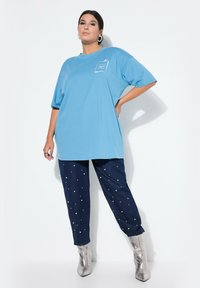 Světle modré oversized tričko z bavlny s krátkými rukávy a bílým potištěným logem. Tmavé džíny s malými kulatými ozdobami. Stříbrné kotníčkové boty.