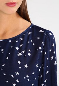 Top blu navy con stampa di stelle bianche, scollo rotondo e tessuto morbido con leggero lucido. Il design include stelle di diverse dimensioni e forme.
