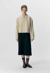 Giacca cropped beige con zip frontale; abbinata a un abito midi nero. La modella indossa mocassini neri e calze trasparenti con un bordo nero.
